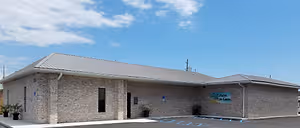 An image of a building with a gray roof and a blue sign that reads All Palm Eye Care, with potted plants in front of it